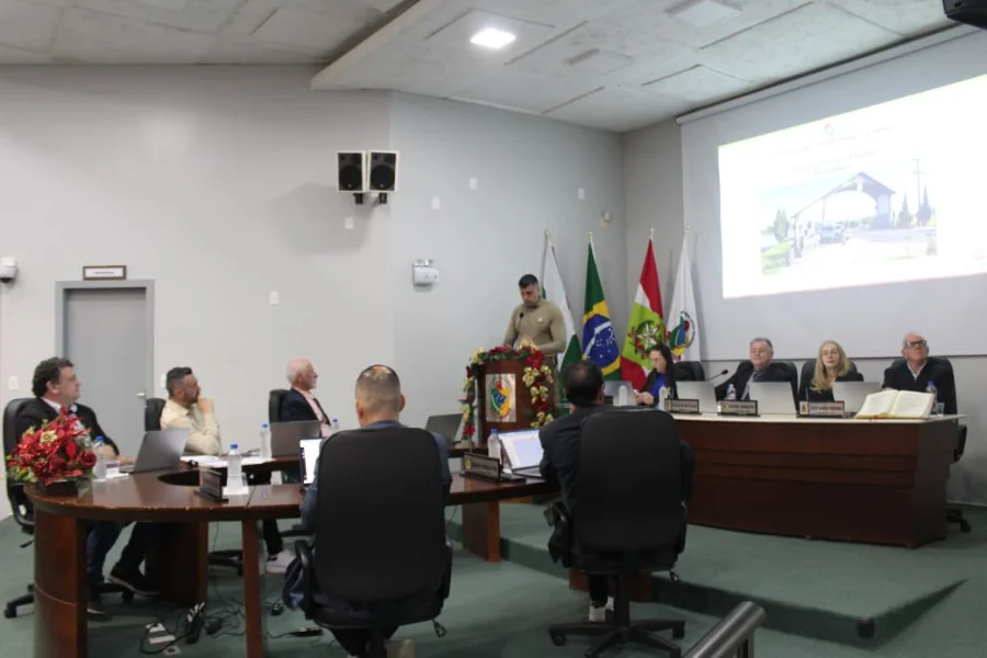 Câmara de Forquilhinha aprova apoio social e ouve balanço da PM Sessão Ordinária na Câmara de Vereadores de Forquilhinha. O Capitão Ramon Marcelina Spillere, de uniforme militar, está em pé em um púlpito de madeira com arranjos de Natal, apresentando o balanço da segurança pública. Os vereadores estão sentados ao redor de uma mesa semicircular em primeiro plano, alguns com notebooks. Ao fundo, uma tela de projeção exibe uma imagem de pórtico da cidade e há quatro bandeiras.