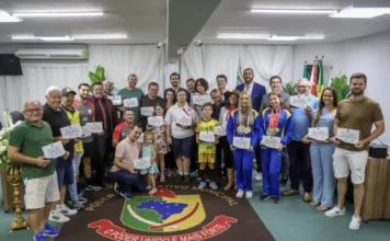 Câmara de Sombrio encerra 2025 com entrega de honrarias Grande grupo de pessoas, incluindo crianças e atletas com medalhas e diplomas, posam para foto oficial no plenário da Câmara de Vereadores de Sombrio, sobre um tapete com o brasão municipal.