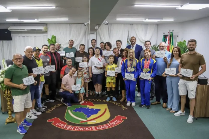 Câmara de Sombrio encerra 2025 com entrega de honrarias Grande grupo de pessoas, incluindo crianças e atletas com medalhas e diplomas, posam para foto oficial no plenário da Câmara de Vereadores de Sombrio, sobre um tapete com o brasão municipal.