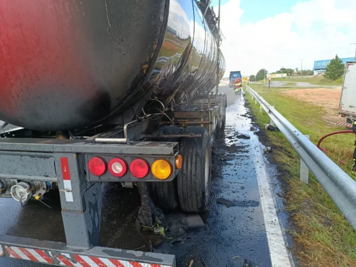 Carreta carregada com xarope de glicose pega fogo na BR-101 Vista traseira de uma carreta tanque preta com pneus queimados e detritos na lateral, após ser atingida por incêndio na BR-101.
