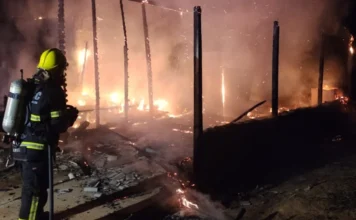 Casa de madeira é destruída por incêndio no município de Ermo Bombeiro de costas, usando capacete amarelo e equipamento de proteção completo, combate incêndio em uma casa de madeira durante a noite. As chamas laranjas intensas consomem a estrutura, restando apenas as vigas carbonizadas. Fumaça densa e faíscas preenchem o ar.