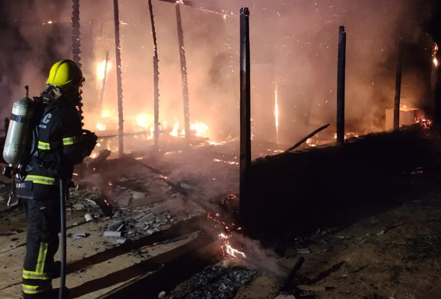 Casa de madeira é destruída por incêndio no município de Ermo Bombeiro de costas, usando capacete amarelo e equipamento de proteção completo, combate incêndio em uma casa de madeira durante a noite. As chamas laranjas intensas consomem a estrutura, restando apenas as vigas carbonizadas. Fumaça densa e faíscas preenchem o ar.