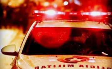 Casal é preso após furtar bolsa durante batizado em Criciúma Fotografia noturna com foco no giroflex vermelho aceso de uma viatura da Polícia Militar de Santa Catarina. O reflexo das luzes vermelhas é visível no capô do veículo, onde se lê a palavra "Polícia" de forma invertida. Ilustra a prisão de casal por furto em Criciúma.