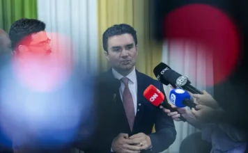 Celso Sabino deixa Ministério do Turismo; União indica sucessor O ministro do Turismo, Celso Sabino, em um pronunciamento oficial. Ele veste um terno escuro, camisa branca e gravata em tom sóbrio. O ministro está posicionado de frente, com uma expressão serena, à frente de um fundo neutro e desfocado que sugere um ambiente de gabinete governamental.