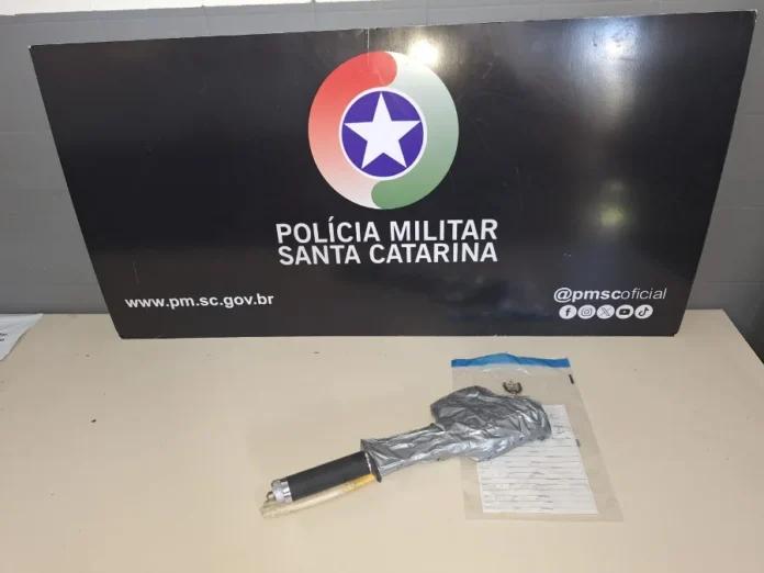 Cocal do Sul Mulher é sequestrada e resgatada pela PM Objeto envolto em fita cinza e um cabo preto (arma improvisada) sobre uma mesa em frente ao banner da Polícia Militar de Santa Catarina.