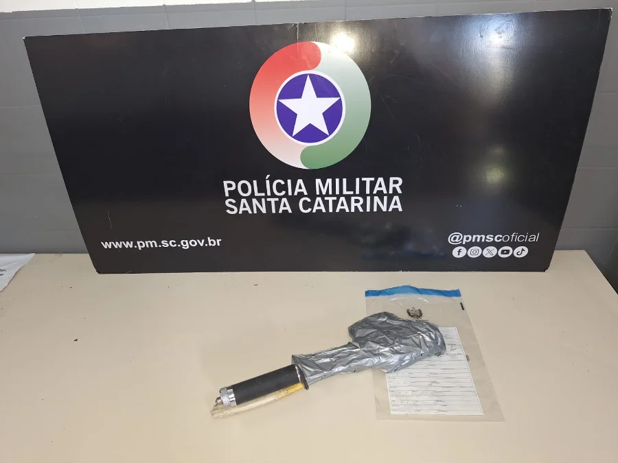 Objeto envolto em fita cinza e um cabo preto (arma improvisada) sobre uma mesa em frente ao banner da Polícia Militar de Santa Catarina.