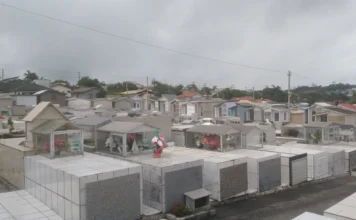 Cocal do Sul abre prazo para regularizar sepulturas Vista panorâmica do Cemitério Municipal de Cocal do Sul sob um céu nublado. Em destaque, diversas sepulturas revestidas de mármore e azulejos brancos, organizadas em fileiras. Ao fundo, casas residenciais da cidade. A imagem ilustra o comunicado da prefeitura sobre o prazo de regularização de titularidade até abril de 2026.