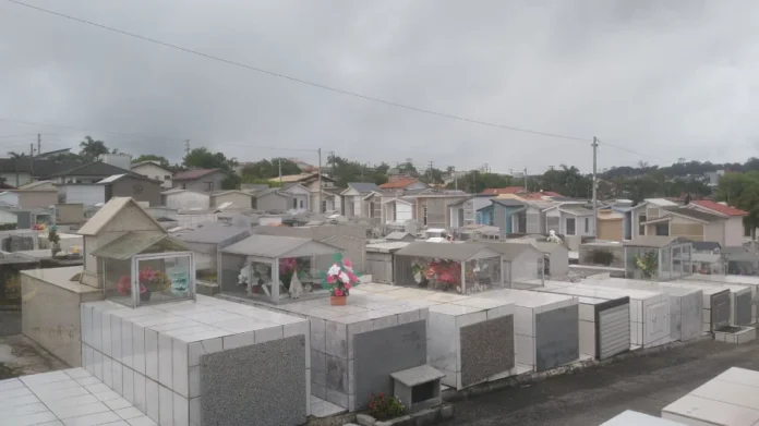 Cocal do Sul abre prazo para regularizar sepulturas Vista panorâmica do Cemitério Municipal de Cocal do Sul sob um céu nublado. Em destaque, diversas sepulturas revestidas de mármore e azulejos brancos, organizadas em fileiras. Ao fundo, casas residenciais da cidade. A imagem ilustra o comunicado da prefeitura sobre o prazo de regularização de titularidade até abril de 2026.
