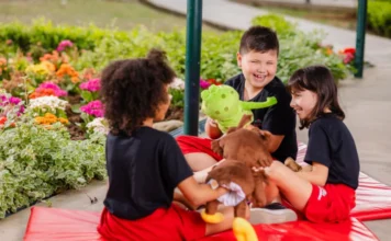 Colégio Unesc abre matrículas para 2026: Integral e Ensino Médio Três crianças, sentadas em um tapete de espuma vermelho ao ar livre, brincam com bichos de pelúcia em um jardim florido. Duas meninas e um menino (este último sorrindo amplamente e segurando um bicho de pelúcia verde) estão interagindo e rindo. O ambiente é cercado por grama e plantas com flores coloridas, ilustrando o ambiente do Colégio Unesc que abriu matrículas para 2026.