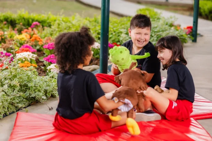 Colégio Unesc abre matrículas para 2026 Integral e Ensino Médio Três crianças, sentadas em um tapete de espuma vermelho ao ar livre, brincam com bichos de pelúcia em um jardim florido. Duas meninas e um menino (este último sorrindo amplamente e segurando um bicho de pelúcia verde) estão interagindo e rindo. O ambiente é cercado por grama e plantas com flores coloridas, ilustrando o ambiente do Colégio Unesc que abriu matrículas para 2026.