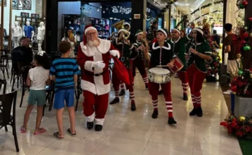 Comércio de Araranguá atende até às 22h nesta reta final Papai Noel caminha pelo corredor de um shopping ou centro comercial em Araranguá, acompanhado por uma banda de ajudantes vestidos de elfos tocando instrumentos de sopro e percussão. Crianças observam a passagem do grupo. A imagem destaca a reta final de vendas de Natal com horário ampliado até as 22h.
