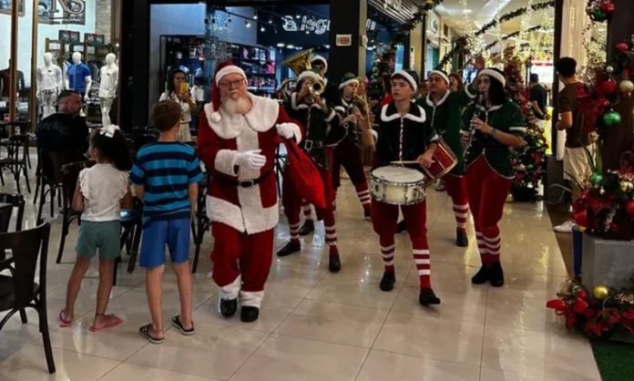 Comércio de Araranguá atende até às 22h nesta reta final Papai Noel caminha pelo corredor de um shopping ou centro comercial em Araranguá, acompanhado por uma banda de ajudantes vestidos de elfos tocando instrumentos de sopro e percussão. Crianças observam a passagem do grupo. A imagem destaca a reta final de vendas de Natal com horário ampliado até as 22h.