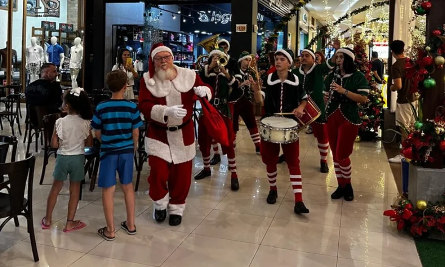 Papai Noel caminha pelo corredor de um shopping ou centro comercial em Araranguá, acompanhado por uma banda de ajudantes vestidos de elfos tocando instrumentos de sopro e percussão. Crianças observam a passagem do grupo. A imagem destaca a reta final de vendas de Natal com horário ampliado até as 22h.