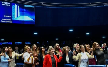 Congresso aprova 19 leis de proteção à mulher em 2025 Um grupo de cerca de quinze deputadas e senadoras está reunido no plenário do Senado Federal, sorrindo e de mãos dadas em sinal de vitória. Ao fundo, um grande painel eletrônico azul exibe "VOTAÇÃO EM ANDAMENTO" e o quorum da sessão. A imagem ilustra o recorde de 19 leis de proteção à mulher aprovadas em 2025.