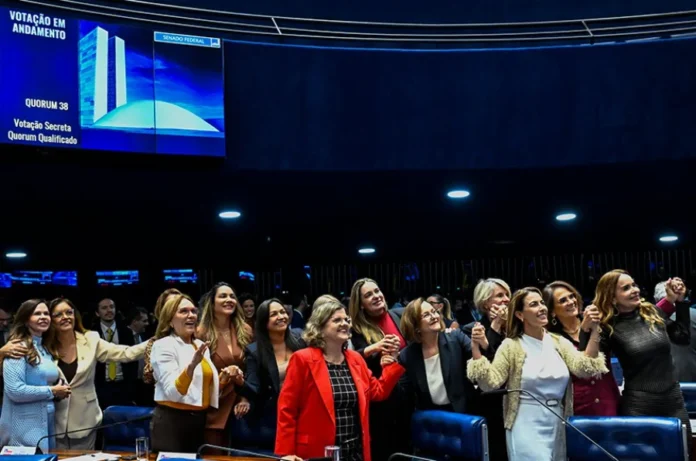 Congresso aprova 19 leis de proteção à mulher em 2025 Um grupo de cerca de quinze deputadas e senadoras está reunido no plenário do Senado Federal, sorrindo e de mãos dadas em sinal de vitória. Ao fundo, um grande painel eletrônico azul exibe "VOTAÇÃO EM ANDAMENTO" e o quorum da sessão. A imagem ilustra o recorde de 19 leis de proteção à mulher aprovadas em 2025.