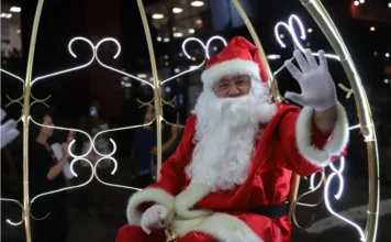Criciúma: Coral encerra programação de Natal nesta quarta Homem vestido de Papai Noel acena com uma luva branca enquanto segura um sino dourado, sentado em uma carruagem iluminada por luzes brancas em formato de arabescos durante um evento noturno.