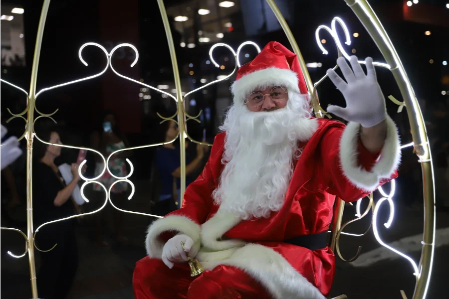 Homem vestido de Papai Noel acena com uma luva branca enquanto segura um sino dourado, sentado em uma carruagem iluminada por luzes brancas em formato de arabescos durante um evento noturno.