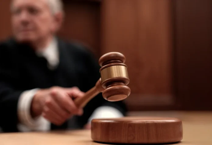 Criciúma Homem é condenado a 41 anos por tirar a vida da ex Imagem de close-up, em foco raso, de uma mão segurando um martelo de juiz (malhete) de madeira com anéis de metal dourado, prestes a bater na base de madeira que o acompanha. No fundo desfocado, há um homem de terno preto e gola branca (possivelmente um juiz), sentado em uma sala de audiências ou tribunal com painéis de madeira. A foto ilustra a sentença de condenação do feminicida pelo Tribunal do Júri de Criciúma.