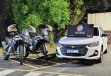 Criciúma: Homem é preso após fuga em carro roubado e clonado Fotografia noturna mostrando um Chevrolet Onix branco estacionado ao lado de duas motocicletas da Polícia Militar de Santa Catarina. Sobre o capô do carro, há um banner preto com o brasão da corporação. Ilustra a recuperação de veículo clonado após perseguição.