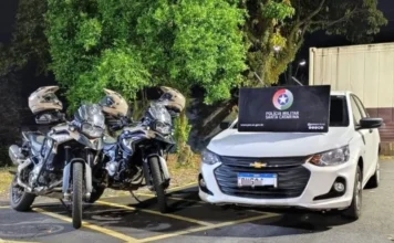 Criciúma: Homem é preso após fuga em carro roubado e clonado Fotografia noturna mostrando um Chevrolet Onix branco estacionado ao lado de duas motocicletas da Polícia Militar de Santa Catarina. Sobre o capô do carro, há um banner preto com o brasão da corporação. Ilustra a recuperação de veículo clonado após perseguição.