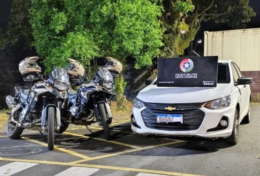 Fotografia noturna mostrando um Chevrolet Onix branco estacionado ao lado de duas motocicletas da Polícia Militar de Santa Catarina. Sobre o capô do carro, há um banner preto com o brasão da corporação. Ilustra a recuperação de veículo clonado após perseguição.