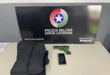 Criciúma: Homem é preso após roubar celular com arma falsa Jaqueta preta, simulacro de pistola verde e um celular sobre uma mesa, em frente ao painel da Polícia Militar de Santa Catarina.