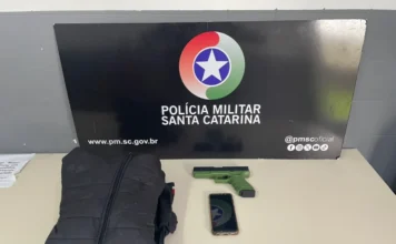 Criciúma: Homem é preso após roubar celular com arma falsa Jaqueta preta, simulacro de pistola verde e um celular sobre uma mesa, em frente ao painel da Polícia Militar de Santa Catarina.