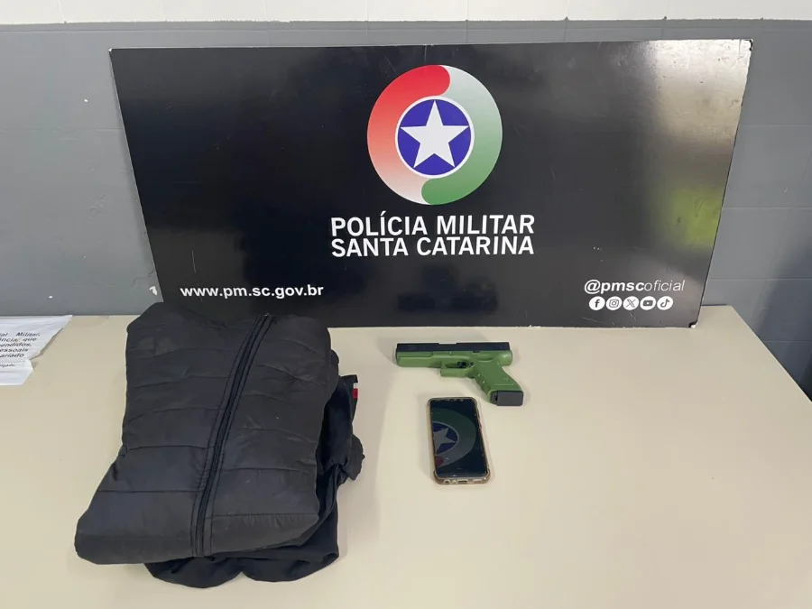 Criciúma Homem é preso após roubar celular com arma falsa Jaqueta preta, simulacro de pistola verde e um celular sobre uma mesa, em frente ao painel da Polícia Militar de Santa Catarina.