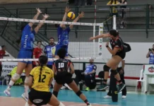Criciúma: Mampituba/Unesc estreia na Superliga B com derrota Cena de jogo de voleibol feminino. No centro, duas jogadoras do time com camisa azul (Mampituba/Unesc) bloqueiam uma bola sobre a rede. Uma jogadora do time adversário, vestindo preto e vermelho com shorts, salta para cortar a bola por cima do bloqueio. Em primeiro plano, uma jogadora de camisa amarela e preta (com o número 12) se prepara para a defesa. A imagem registra um momento da partida de estreia do Mampituba/FME Criciúma/Radar/Unesc na Superliga B contra o Abel Moda Vôlei.