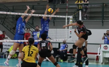 Criciúma: Mampituba/Unesc estreia na Superliga B com derrota Cena de jogo de voleibol feminino. No centro, duas jogadoras do time com camisa azul (Mampituba/Unesc) bloqueiam uma bola sobre a rede. Uma jogadora do time adversário, vestindo preto e vermelho com shorts, salta para cortar a bola por cima do bloqueio. Em primeiro plano, uma jogadora de camisa amarela e preta (com o número 12) se prepara para a defesa. A imagem registra um momento da partida de estreia do Mampituba/FME Criciúma/Radar/Unesc na Superliga B contra o Abel Moda Vôlei.