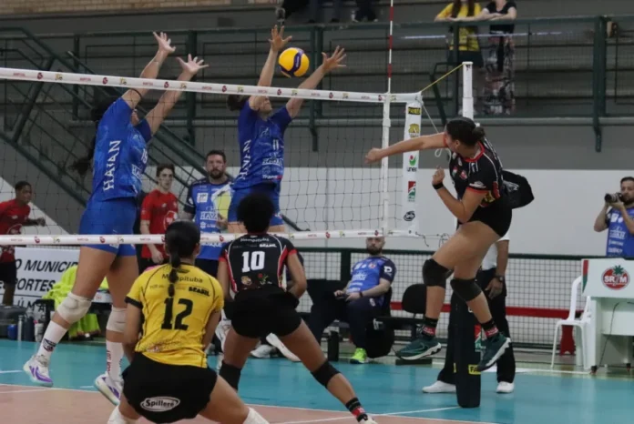 Criciúma Mampituba Unesc estreia na Superliga B com derrota Cena de jogo de voleibol feminino. No centro, duas jogadoras do time com camisa azul (Mampituba/Unesc) bloqueiam uma bola sobre a rede. Uma jogadora do time adversário, vestindo preto e vermelho com shorts, salta para cortar a bola por cima do bloqueio. Em primeiro plano, uma jogadora de camisa amarela e preta (com o número 12) se prepara para a defesa. A imagem registra um momento da partida de estreia do Mampituba/FME Criciúma/Radar/Unesc na Superliga B contra o Abel Moda Vôlei.