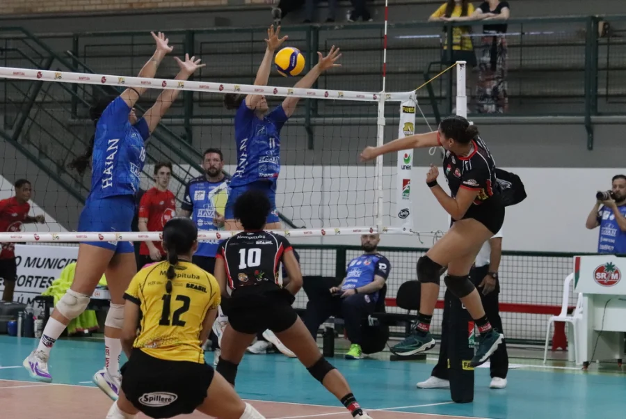 Criciúma Mampituba Unesc estreia na Superliga B com derrota Cena de jogo de voleibol feminino. No centro, duas jogadoras do time com camisa azul (Mampituba/Unesc) bloqueiam uma bola sobre a rede. Uma jogadora do time adversário, vestindo preto e vermelho com shorts, salta para cortar a bola por cima do bloqueio. Em primeiro plano, uma jogadora de camisa amarela e preta (com o número 12) se prepara para a defesa. A imagem registra um momento da partida de estreia do Mampituba/FME Criciúma/Radar/Unesc na Superliga B contra o Abel Moda Vôlei.