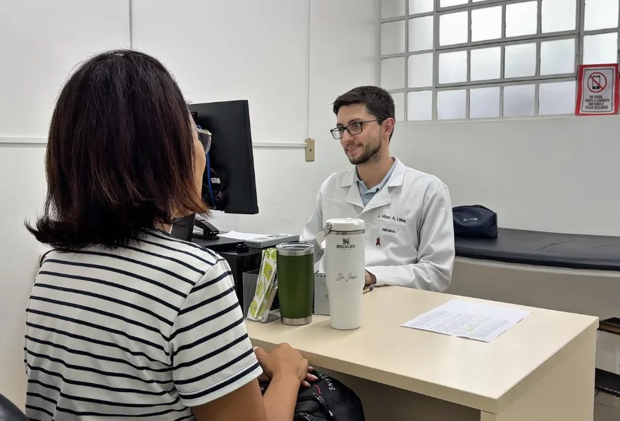 Médico jovem de jaleco branco, sorridente, atende uma paciente de costas em um consultório. Sobre a mesa, há um monitor, um copo térmico branco e papéis. Ao fundo, uma maca e uma janela com grade.