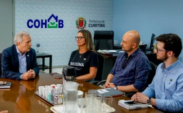 Criciúma busca Curitiba para modelos de habitação popular Reunião de trabalho na Companhia de Habitação Popular de Curitiba (Cohab). Quatro pessoas estão sentadas ao redor de uma mesa de madeira escura com um prato central com copos e jarras. O presidente da Cohab, à esquerda, fala com os três gestores de Criciúma. Ao fundo, nas paredes azuis, estão as logomarcas 'COHAB' e 'Prefeitura de Curitiba'.