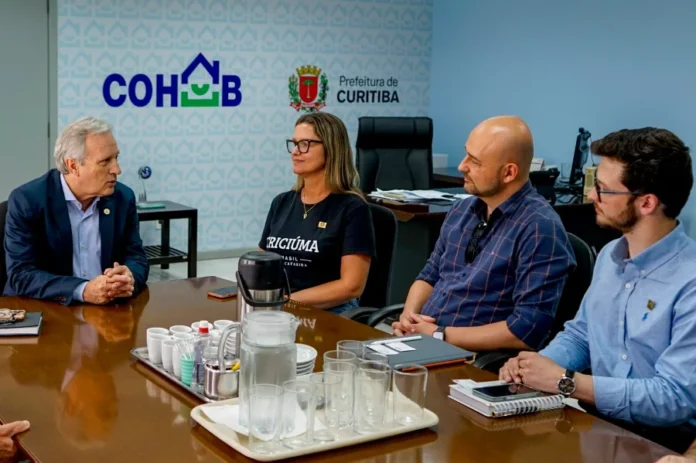 Criciúma busca Curitiba para modelos de habitação popular Reunião de trabalho na Companhia de Habitação Popular de Curitiba (Cohab). Quatro pessoas estão sentadas ao redor de uma mesa de madeira escura com um prato central com copos e jarras. O presidente da Cohab, à esquerda, fala com os três gestores de Criciúma. Ao fundo, nas paredes azuis, estão as logomarcas 'COHAB' e 'Prefeitura de Curitiba'.