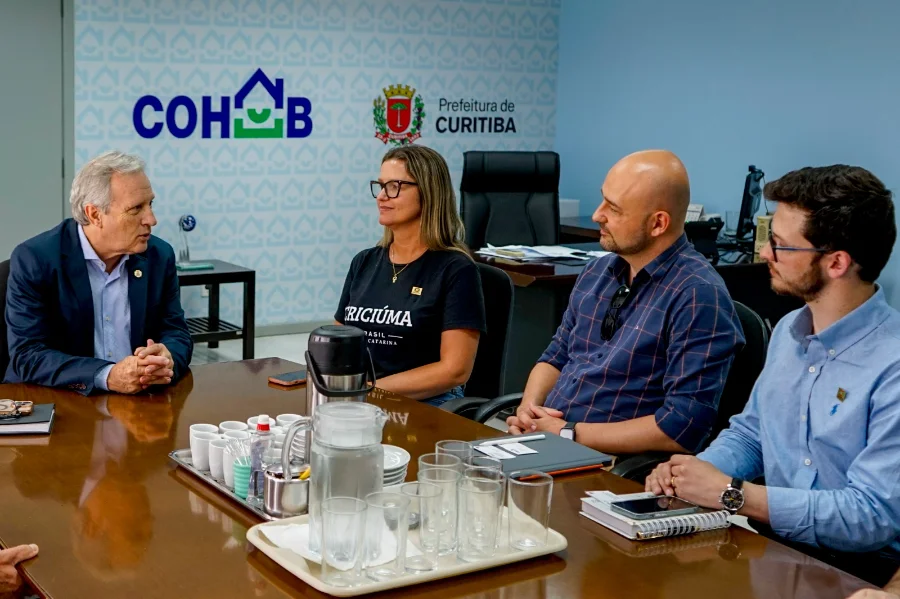 Criciúma busca Curitiba para modelos de habitação popular Reunião de trabalho na Companhia de Habitação Popular de Curitiba (Cohab). Quatro pessoas estão sentadas ao redor de uma mesa de madeira escura com um prato central com copos e jarras. O presidente da Cohab, à esquerda, fala com os três gestores de Criciúma. Ao fundo, nas paredes azuis, estão as logomarcas 'COHAB' e 'Prefeitura de Curitiba'.