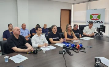 Criciúma cria “Central do Recomeço” contra dependência química "Prefeito Vagner Espíndola (no centro, de camisa preta com a inscrição 'Criciúma Brasil') e outros secretários municipais e autoridades sentados em uma longa mesa de reunião, em frente a microfones de imprensa. Estão presentes os secretários Thiago Fabris e Dudi Sônego e outros gestores. Ao fundo, um banner exibe o logo 'Criciúma: O Trabalho Gera o Futuro'."