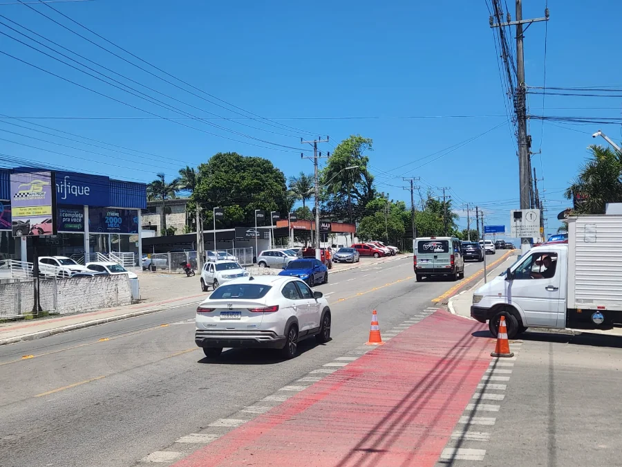 Carros trafegando em uma avenida (Luiz Lazzarin) em Criciúma, com a trincheira recém-liberada à frente, sob forte sol e céu azul.