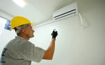 Estado investe R$ 1,1 mi em ar-condicionado para hospitais Um técnico usando capacete de segurança amarelo e luvas pretas utiliza um controle remoto para testar um aparelho de ar-condicionado branco recém-instalado em uma parede clara. A imagem ilustra o investimento de R$ 1,1 milhão do Governo de SC na climatização de hospitais.