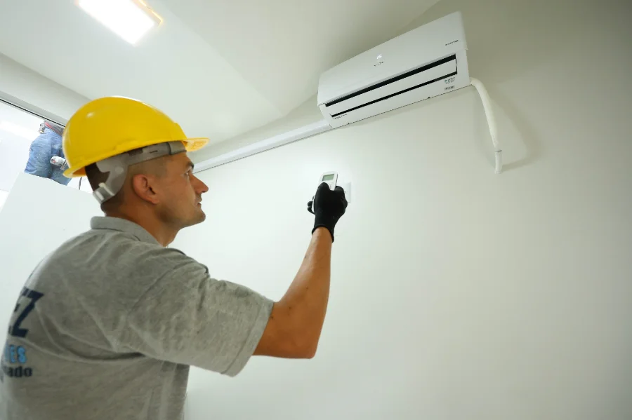 Um técnico usando capacete de segurança amarelo e luvas pretas utiliza um controle remoto para testar um aparelho de ar-condicionado branco recém-instalado em uma parede clara. A imagem ilustra o investimento de R$ 1,1 milhão do Governo de SC na climatização de hospitais.
