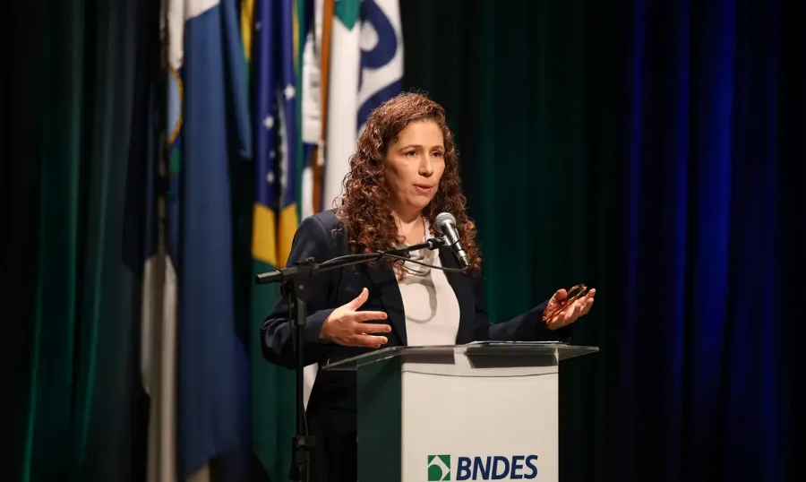 Estatais não são peso e sim ativo do Brasil, diz Esther Dweck A ministra da Gestão e da Inovação em Serviços Públicos, Esther Dweck, em pé, discursa em um púlpito transparente com a logomarca do BNDES. Ela veste terno azul-marinho sobre camisa branca e segura óculos de aro escuro em sua mão direita, gesticulando com a mão esquerda. Ao fundo, estão bandeiras do Brasil e um painel verde escuro.