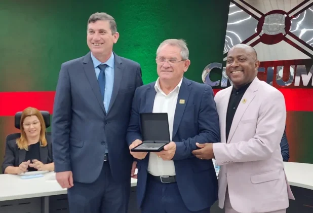 O presidente da C&acirc;mara de Crici&uacute;ma, Marcio Daros (&agrave; esquerda), entrega uma placa de homenagem ao vice-prefeito Sal&eacute;sio Lima (&agrave; direita). Ambos seguram o estojo da placa e sorriem para a foto.