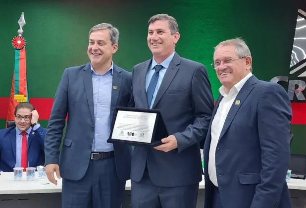 O prefeito Vagner Espindola (&agrave; esquerda) entrega uma placa comemorativa ao presidente da C&acirc;mara, Marcio Daros (&agrave; direita), no Sal&atilde;o Ouro Negro. O ato simboliza o reconhecimento do Executivo ao trabalho do Legislativo no ano do Centen&aacute;rio. Eles est&atilde;o posicionados &agrave; frente de um painel verde com a identidade visual da prefeitura.