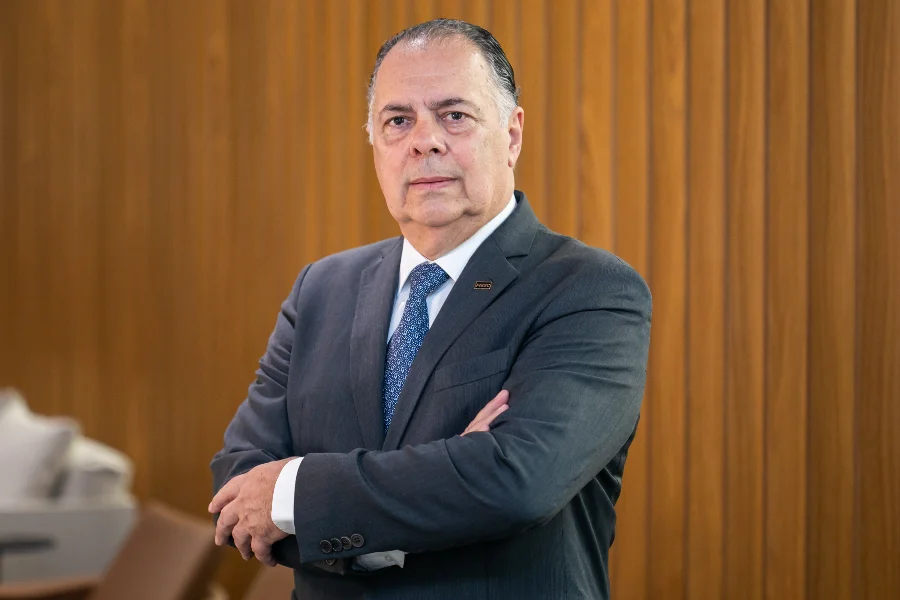 Retrato do presidente da FIESC, Gilberto Seleme, de braços cruzados, vestindo terno cinza e gravata azul. Ele está posicionado em frente a uma parede de painéis de madeira vertical.