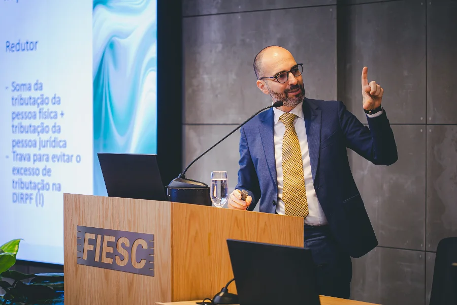 FIESC Isenção do IR traz insegurança jurídica em dividendos O advogado tributarista Vicente Lisboa Capella, de barba e terno azul, gesticula com o dedo indicador esquerdo enquanto profere uma palestra em reunião da FIESC. Ele está em pé em um púlpito de madeira com o logo da Federação. À esquerda, uma tela exibe slides com tópicos de tributação. Há um copo d'água e notebooks sobre a mesa.