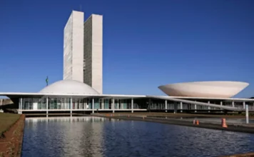 FIESC critica corte de incentivos e aumento de impostos Fotografia externa do Palácio do Congresso Nacional em Brasília sob um céu azul limpo. Em destaque, as duas cúpulas (Câmara e Senado) e as torres gêmeas refletidas no espelho d'água em primeiro plano. A imagem ilustra a crítica da FIESC contra a aprovação do PLP 128/2025 pelo Legislativo.