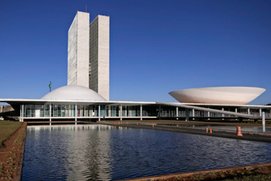 FIESC critica corte de incentivos e aumento de impostos Fotografia externa do Palácio do Congresso Nacional em Brasília sob um céu azul limpo. Em destaque, as duas cúpulas (Câmara e Senado) e as torres gêmeas refletidas no espelho d'água em primeiro plano. A imagem ilustra a crítica da FIESC contra a aprovação do PLP 128/2025 pelo Legislativo.