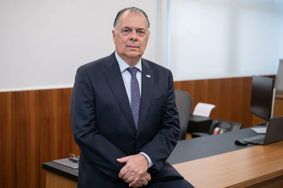 FIESC defende modernização urgente do Mercosul engessado O presidente da FIESC, Gilberto Seleme, em um retrato formal no escritório, vestindo terno azul-marinho e gravata. Ele está sentado na beira de uma mesa de trabalho de madeira escura. Ao fundo, uma parede branca e clean e um monitor de computador.