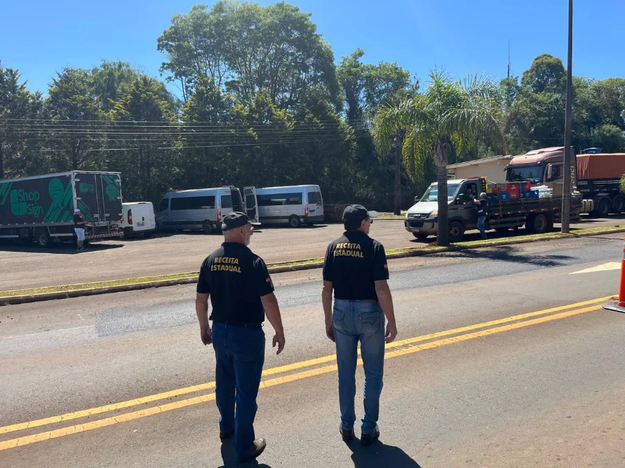 Dois auditores fiscais da Receita Estadual (SC), identificados com camisetas pretas, caminham em uma via, inspecionando caminhões e vans em segundo plano.