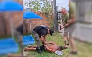 Foragido com pena de 52 anos é preso em Araranguá Foto de uma abordagem policial em um gramado residencial. Um policial civil, usando colete balístico e uniforme preto, algema um homem deitado de bruços no chão. Um segundo policial está em pé ao lado, dando cobertura à ação. Ao fundo, vê-se um guarda-sol azul e uma pequena piscina infantil. O detido é um foragido condenado a 52 anos de prisão.
